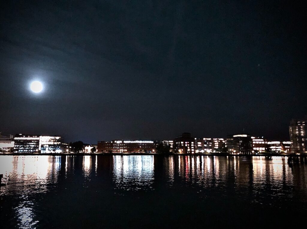 View over the water and the row of houses further away at night. The houses are illuminated, or rather they radiate light which is reflected in the water. The full moon can be seen in the centre of the picture on the left.